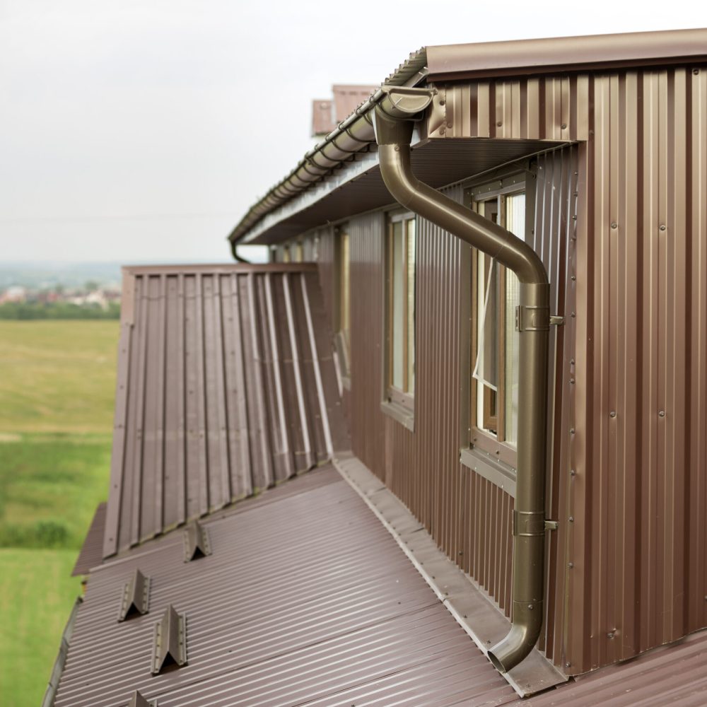 Close-up exterior detail of residential cottage attic room with plastic windows, roof and walls covered with brown metal decorative siding planks, new gutter system on blurred copy space background.